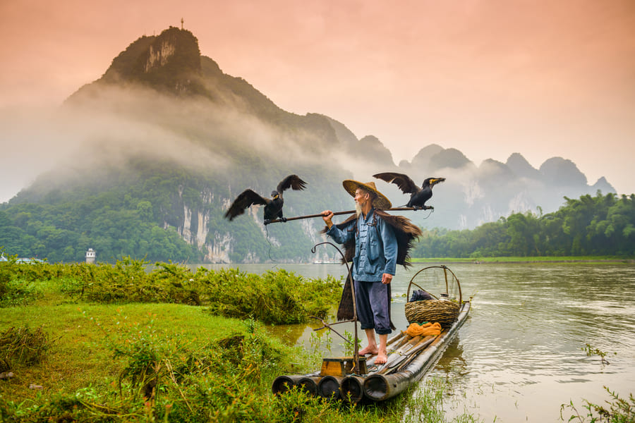 pescador de cormoran con dos aves a los costados en una balsa sobre rio